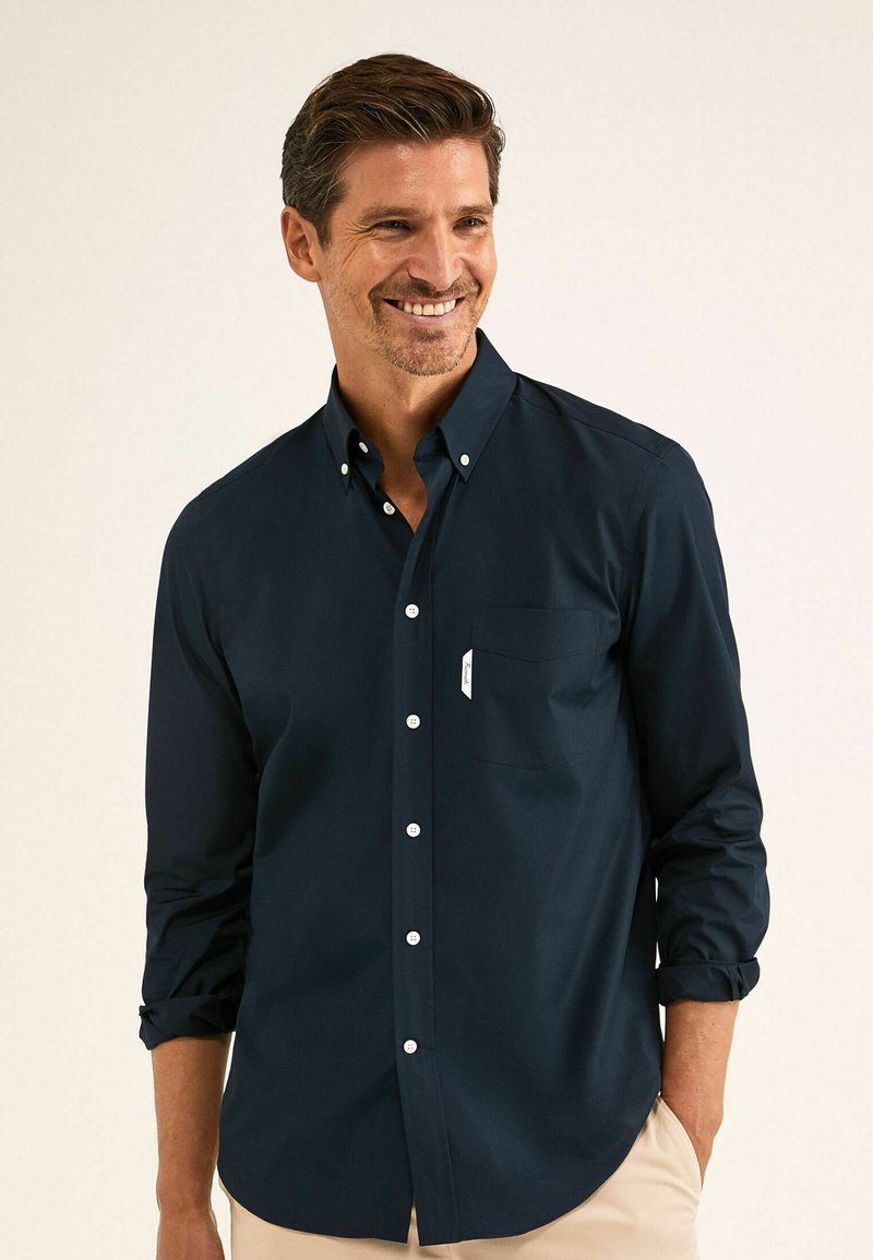 Camicia blu navy con colletto strutturato, maniche lunghe arrotolate ai polsi, tasca sul petto e bottoni bianchi. Tessuto dalla trama liscia.