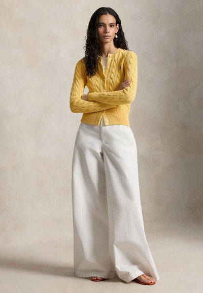 Femeie cu păr lung și creț poartă un cardigan galben tricotat cu model în relief, pantaloni albi largi și sandale portocalii, stând cu brațele încrucișate pe un fundal bej.