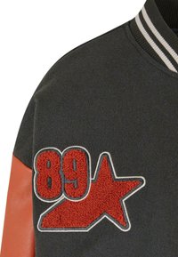Grå ulljakke med oransje skinnærmer, med et strukturert rødt emblem med "89" og en stjerne. Stripete krage gir ekstra detaljer.