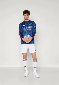 Maillot de gardien de but à manches longues bleu avec un motif géométrique, des accents rouges, un short blanc et des chaussettes assorties, tenant un ballon de football.