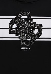 Svart hettegenser med et stort, utsmykket logo med rhinestones, hvite striper, og teksten "GUESS EST.1981" nederst.