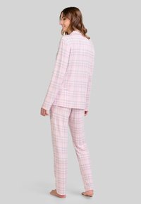 Mujer de pie descalza, de perfil, vistiendo pijamas de manga larga de cuadros rosa y gris con pantalones a juego, sobre un fondo gris liso.