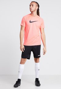 Nike neon rožinė sportinė marškinė su juodu logotipu, derinama su juodomis šortais ir baltais iki kelių kojinėmis. Avalynė - juodi futbolo batai.