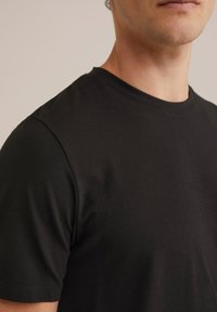 Primer plano de una persona que lleva una camiseta negra de manga corta y cuello redondo sobre un fondo neutro.