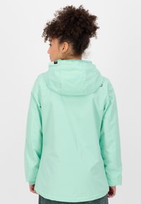 Veste imperméable vert menthe avec capuche, coupe ample et texture lisse. Présente une étiquette logo sur le col et des poignets élastiques.