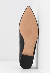 Chaussure en cuir noir à bout pointu reposant sur le côté, montrant une semelle beige avec de fines lignes diagonales antidérapantes et un petit talon noir.