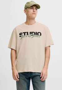 Beżowa koszulka typu t-shirt o luźnym kroju, z czarnym i zielonym nadrukiem tekstu "STUDIO Jack & Jones". W połączeniu z niebieskimi dżinsami. Prosty design, bawełnowy materiał.