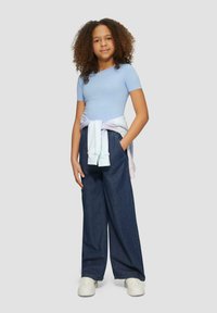 Top claro azul de manga corta acanalada, pantalones anchos de mezclilla oscuros y holgados, sudadera en tonos pastel atada a la cintura, zapatillas blancas, cabello rizado.