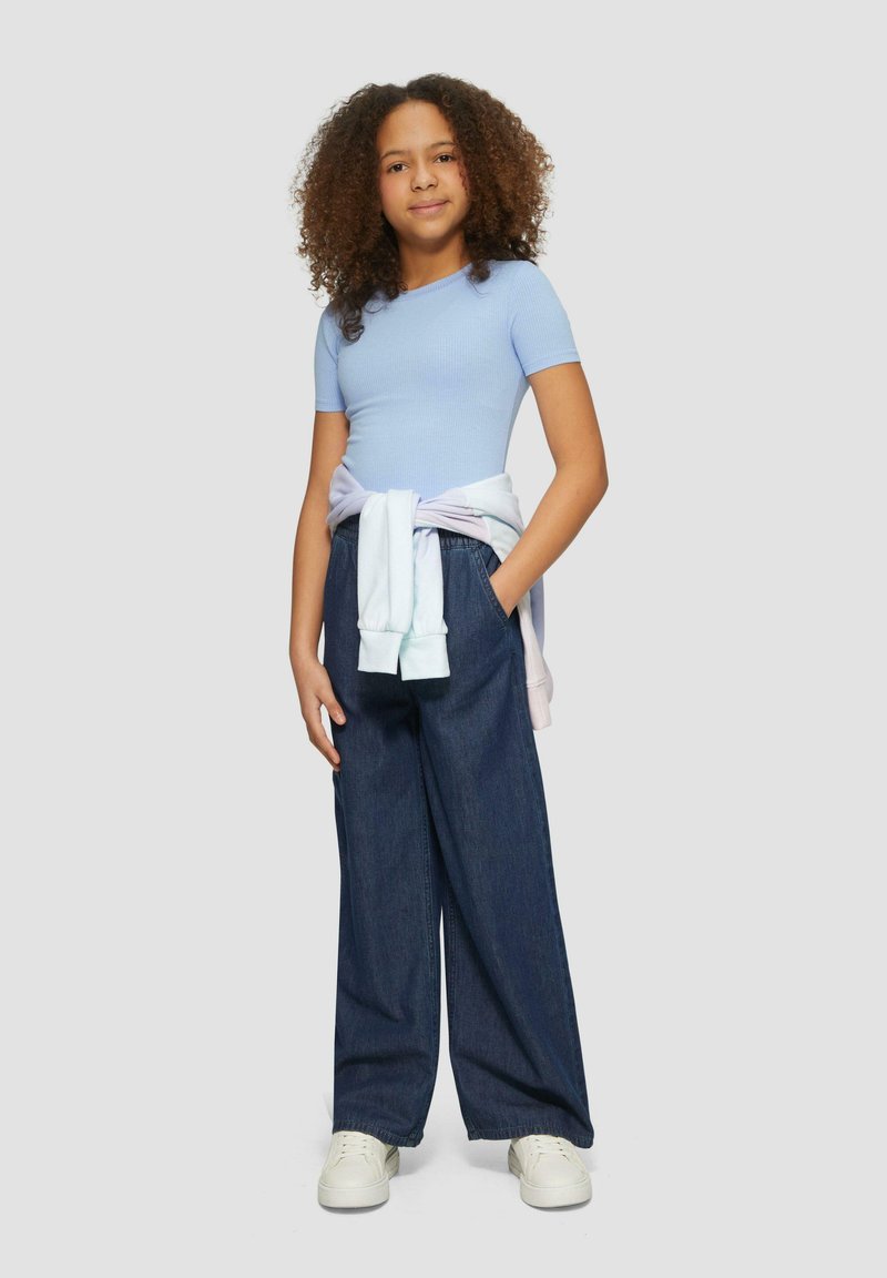Top claro azul de manga corta acanalada, pantalones anchos de mezclilla oscuros y holgados, sudadera en tonos pastel atada a la cintura, zapatillas blancas, cabello rizado.