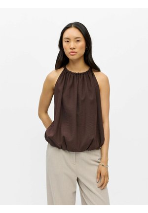 Femme aux longs cheveux noirs portant un haut dos nu brun foncé froncé et un pantalon beige clair, debout devant un fond blanc uni.