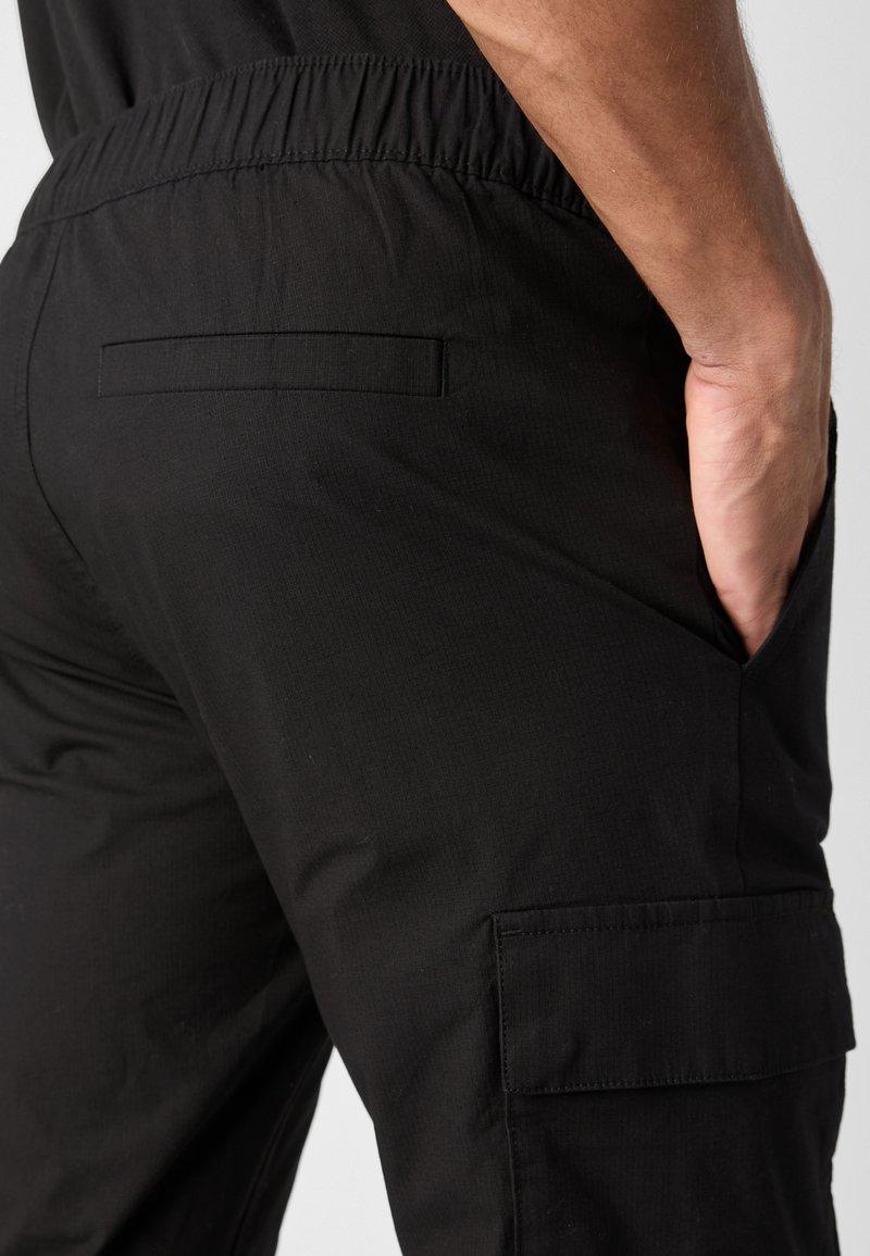 Pantaloni cargo neri con cintura elasticizzata, tasche laterali e una tasca sul retro. Il materiale sembra liscio con una leggera texura.