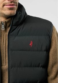 Čierna puffová vesta s vysokým golierom, ktorá má na ľavej strane hrudníka vyšívané červené logo. Materiál vyzerá hladko a izolačne.