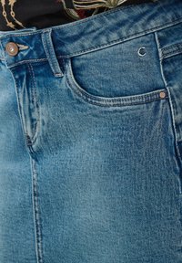 Gros plan d'une poche de jean en denim bleu avec rivets en cuivre, passant de ceinture et fermeture par bouton, porté avec une chemise à motif floral.