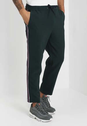 Uomo che indossa pantaloni jogger verde scuro con strisce laterali e scarpe da ginnastica grigie, in piedi con le mani in tasca davanti a uno sfondo semplice.