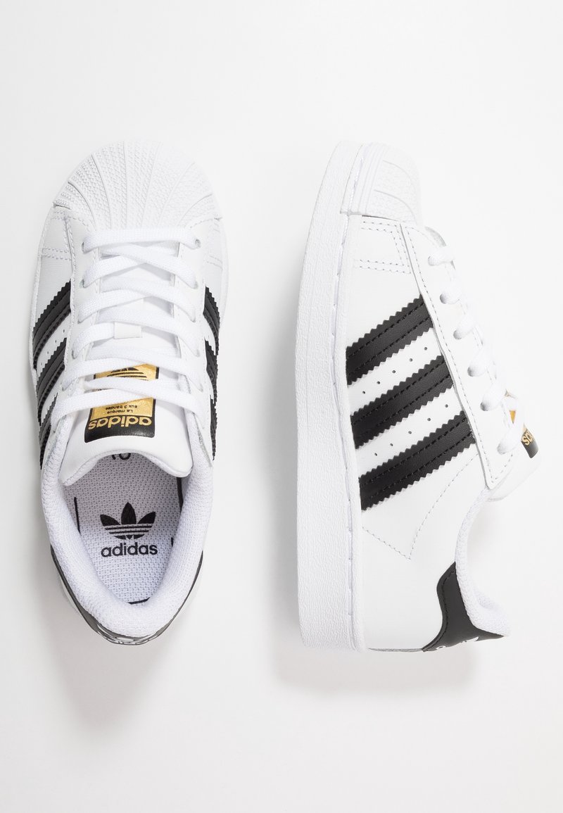 Valkoiset Adidas-tennarit mustilla raidoilla ja korosteväreillä, joissa on nauhat ja kultainen logo kielekkeessä, esitelty ylhäältäpäin.