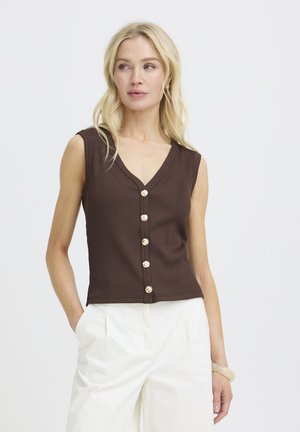 Kvinde med langt blondt hår iført brun, ærmeløs knapbluse og hvide bukser, stående med den ene hånd i lommen.