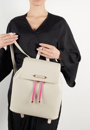 Beżowy skórzany plecak z klapą, z złotymi elementami i jasnoróżowymi sznurkami. Gładka tekstura z wyraźnie zdefiniowaną formą.