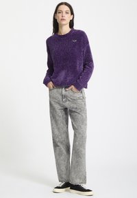 Lilla tekstureret sweater med rund hals, parret med grå vide bukser og sorte sneakers med hvide såler. Diskret branding på sweateren.