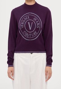 Pull en maille violet avec un logo circulaire Versace Jeans Couture argenté, doté d'un col et de poignets côtelés. Assorti à un pantalon rayé.
