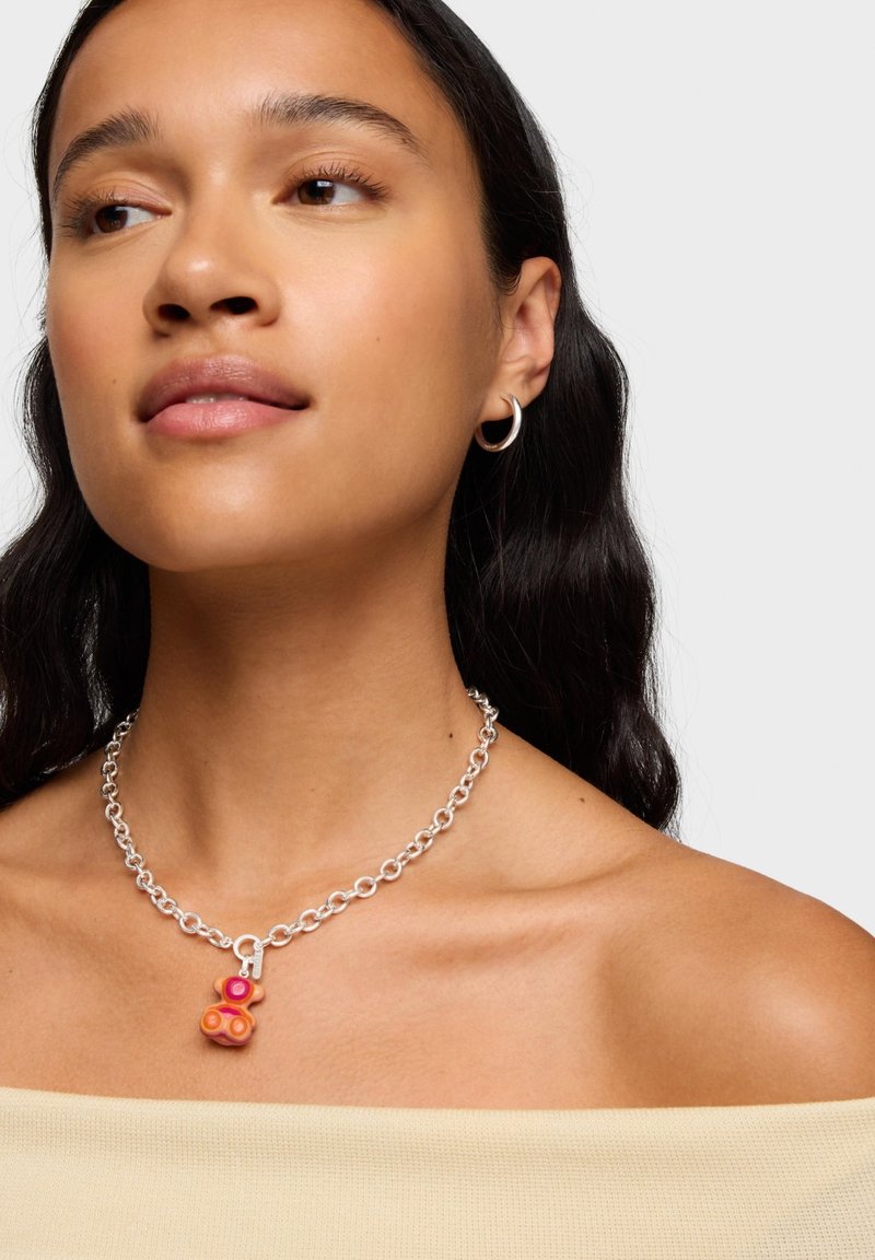 Collana con catena d'argento con ciondolo a forma di orsetto multicolore con accenti rosa e arancioni. Texture liscia e maglie circolari sono evidenti.