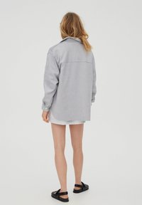 Grå oversized skjorte lavet af blød stof, der har lange ærmer, en spids krave og en afrundet kant, parret med grå shorts og sorte sandaler.