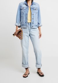 Veste en denim délavé avec deux poches poitrine, associée à un haut jaune et un jean taille haute en denim clair, complétée par des sandales noires à lanières.