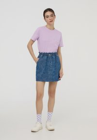 T-shirt lilla con taschino sul davanti, abbinato a una gonna di jeans blu con un motivo disegnato a mano, e calze a quadri viola. Sneakers bianche.