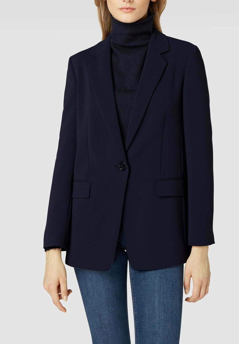 Femme portant un blazer noir par-dessus un col roulé noir et un jean bleu, debout devant un fond gris clair uni.