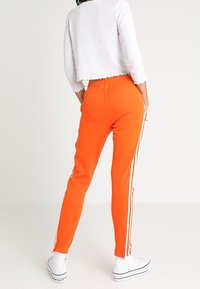 Henkilö kirkkaan oransseissa treenihousuissa, joissa on valkoiset sivuraidat, valkoisessa lyhyessä pitkähihassa paidassa ja valkoisissa korkeissa tennareissa, seisoo tasaisella taustalla.