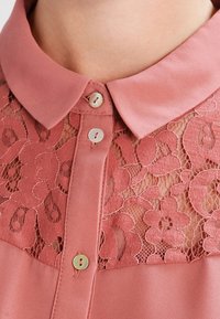 Růžová blůza z jemného materiálu, s krajkovou panelou s květinovými vzory, zapínáním na knoflíky a límečkem.