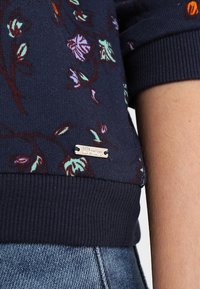 Maglione blu navy con motivo floreale in verde chiaro e viola. Presenta polsini e orlo a coste, etichetta in metallo con decorazione "Denim".