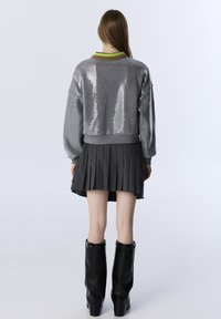 Grijze cropped trui met pailletten, groene en bruine kraag; grijze geplooide rok, zwarte glanzende laarzen tot de knie. Soepele textuur, modern ontwerp.