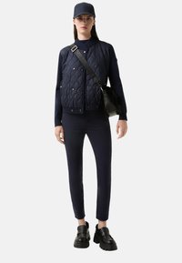 Granatowa pikowana kurtka z srebrnymi zatrzaskami, dopasowane granatowe spodnie oraz czarne buty na platformie. Dodatki obejmują pasującą czapkę i torbę na ramię.