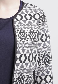 Černobílý cardigan s geometrickými vzory, strukturovaná pletenina, otevřený přední díl a kontrastní jednolitá námořnická modrá podšívka.