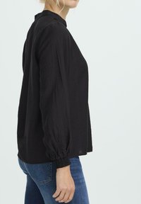 Frau in einer langärmeligen schwarzen Bluse mit gerafften Manschetten und blauen Jeans, von der Seite vor einem schlichten hellen Hintergrund dargestellt.