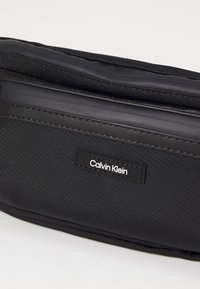 Zwarte textiel pouch met gladde en gestructureerde materialen, een ritsluiting en een 'Calvin Klein' logo label aan de voorkant.
