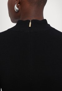 Gros plan sur une personne portant un haut noir côtelé avec une tirette en or au col arrière et une boucle d'oreille argentée en forme de spirale.