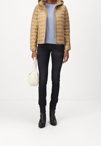 Beżowa pikowana kurtka z kapturem, niebieska bluzka, ciemne dżinsy i czarne botki za kostkę. Trzyma małą białą torebkę.