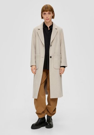 Lange beige wollen jas met gesneden revers, twee voorkeurszakken en zwarte knopen, gedragen over een zwart shirt en losse bruine broeken met zwarte laarzen.