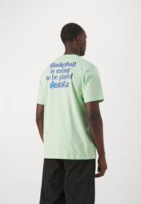 T-shirt in cotone verde chiaro con testo blu sul retro: "Il basket è fatto per essere giocato all'aperto," con una grafica di foglia decorativa.
