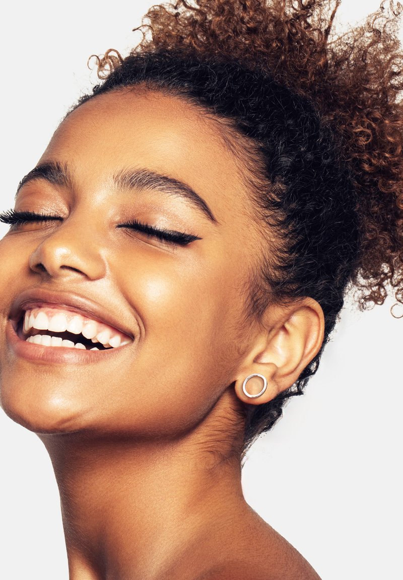 Smiling individual with curly hair, wearing a silver circular earring. Skin is smooth and radiant, with neutral-toned makeup enhancing features.