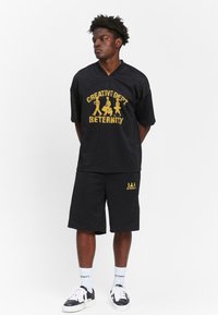 Black mesh short-sleeve shirt with yellow graphic text and accents, paired with matching shorts featuring similar designs; completed with black sneakers.