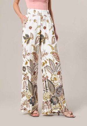 Femme portant un pantalon blanc à jambes larges avec des motifs floraux marron, dorés et noirs, associé à des sandales roses et un haut rose sans manches.
