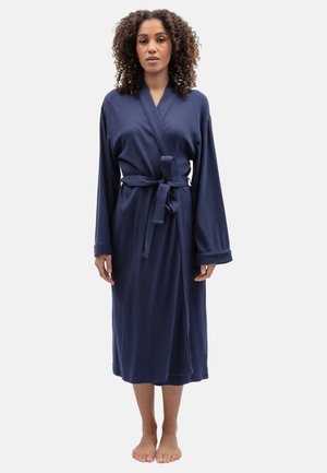 Femme aux cheveux bouclés, debout pieds nus, portant une longue robe bleu marine nouée à la taille sur un fond clair uni.