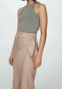 Vrouw in een mouwloze olijfgroene ribgebreide top en een beige wikkelrok die met een strik om de taille is geknoopt, tegen een effen achtergrond.