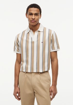 Férfi rövid ujjú, galléros ingben, bézs, fehér és fekete függőleges csíkokkal, valamint bézs nadrágban, egyszínű háttér előtt.