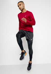 Červené dlouhé rukávy sportovní tričko, černé kraťasy, černé a oranžové běžecké boty, černé strukturované legíny se vzorovanými akcenty, v běžecké pozici.