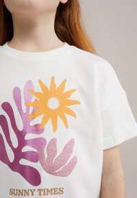 Baltas medvilnės marškinėliai su spalvingais spaudinių: oranžine saule, purpuriniais ir rožiniais lapų dizainais, ir „SUNNY TIMES“ tekstu auksinės spalvos apačioje.