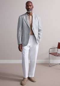 Giacca da completo in lino grigio chiaro sopra una polo a coste beige, abbinata a pantaloni bianchi e mocassini beige. Design semplice, texture liscia, stile formale.