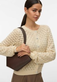Cardigan en tricot beige avec motif ajouré, encolure ronde et boutons décoratifs. Tenant un sac à main minimaliste marron. Pantalon marron avec une texture subtile.